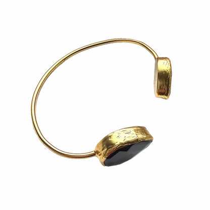 Gold-plated cuff bracelet with two onyx black stones on a white stone surface facing right