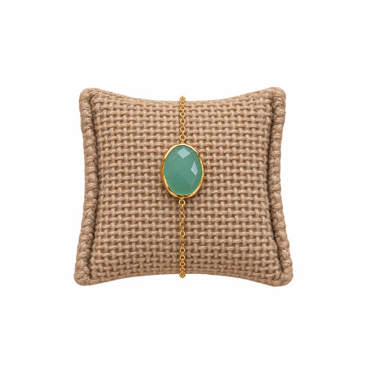 A gold chain bracelet with an oval light green aqua chalcedony gemstone on a woven tan pillow against a white background.