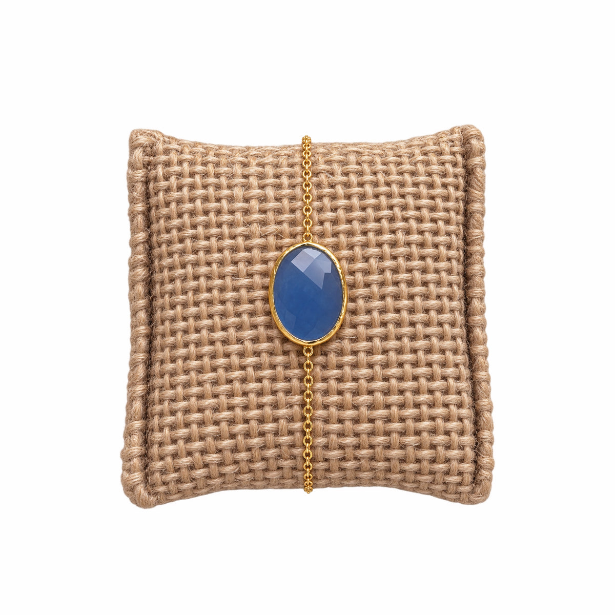 A gold chain bracelet with an oval light blue chalcedony gemstone pendant, displayed on a woven tan jewelry pillow against a white background.