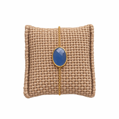 A gold chain bracelet with an oval light blue chalcedony gemstone pendant, displayed on a woven tan jewelry pillow against a white background.