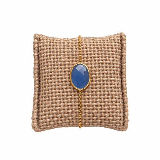 A gold chain bracelet with an oval light blue chalcedony gemstone pendant, displayed on a woven tan jewelry pillow against a white background.