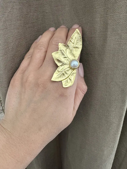 Gold leaf-shaped ring with a central pearl gemstone on a hand against a brown fabric background