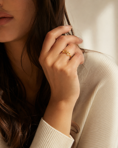 A close-up lifestyle shot of a woman wearing a delicate gold infinity ring on her finger, dressed in a cream ribbed knit sweater against a softly lit, warm-toned background.
