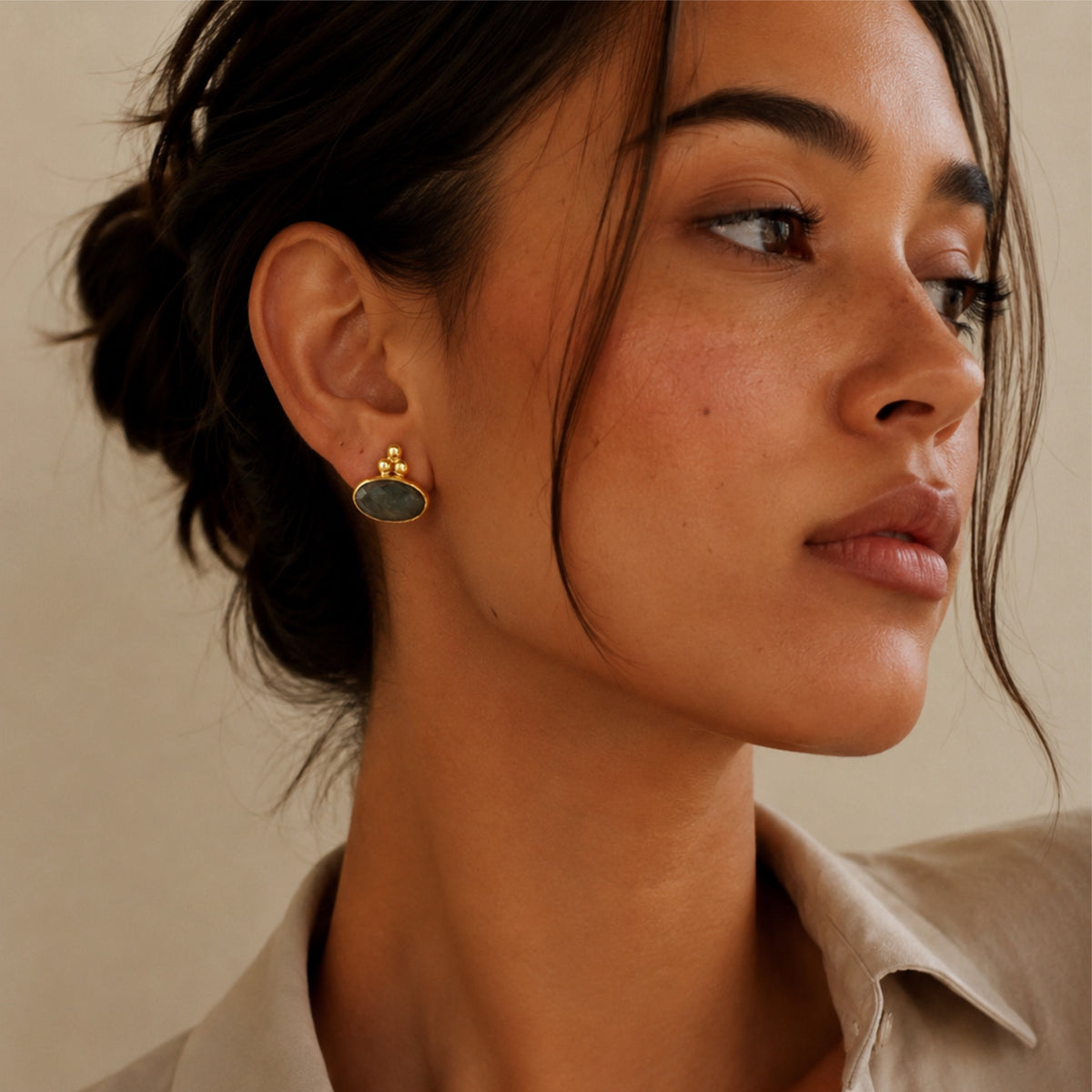 A close-up of a woman wearing a small gold stud earring with an oval labradorite gray stone, shown in profile against a warm beige background.