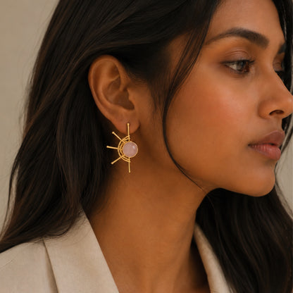 A close-up side profile of a woman wearing a gold sunburst earring with a pale pink rose quartz stone center, paired with a light beige blazer against a soft beige background.