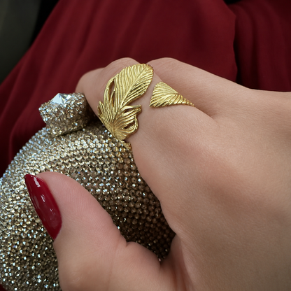 Red manicured nails on hand holding a gold clutch with decorative elements while wearing a gold-plated feather wrap Ring 