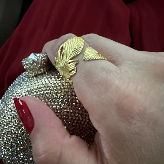 Red manicured nails on hand holding a gold clutch with decorative elements while wearing a gold-plated feather wrap Ring 