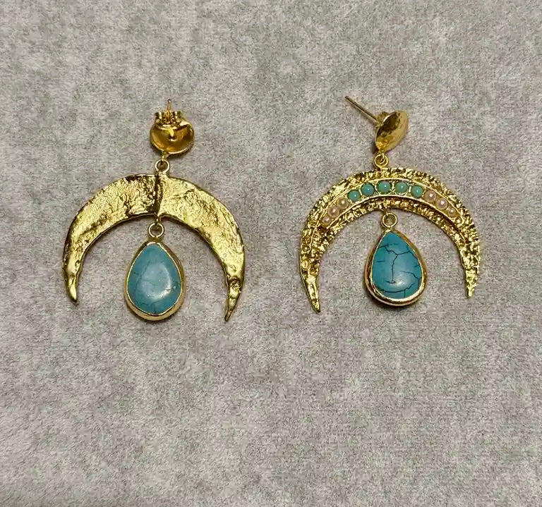 A pair of gold crescent moon drop earrings with pearls and turquoise teardrop stones, displayed on a textured gray background.