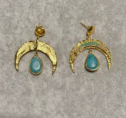A pair of gold crescent moon drop earrings with pearls and turquoise teardrop stones, displayed on a textured gray background.