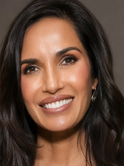 A close-up portrait of Padma Lakshmi wearing a small silver evil eye stud earring, set against a plain taupe-gray background