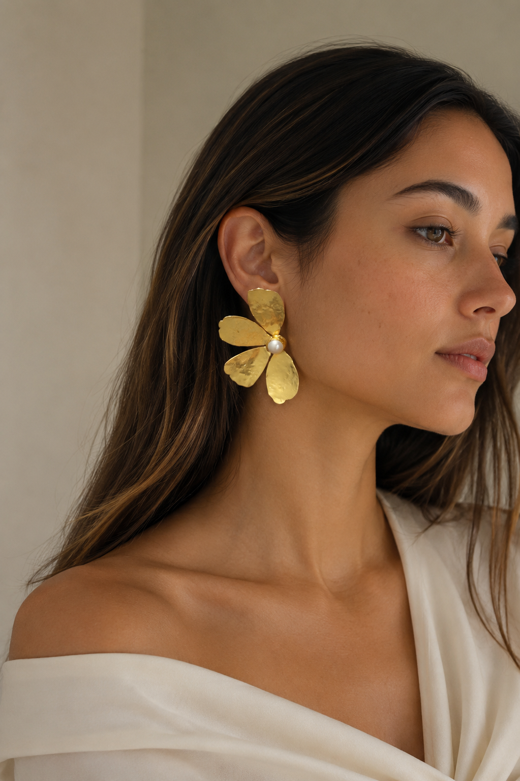 A woman wearing a large, textured gold flower-shaped earring with a small white pearl at the center, paired with a cream off-the-shoulder top.
