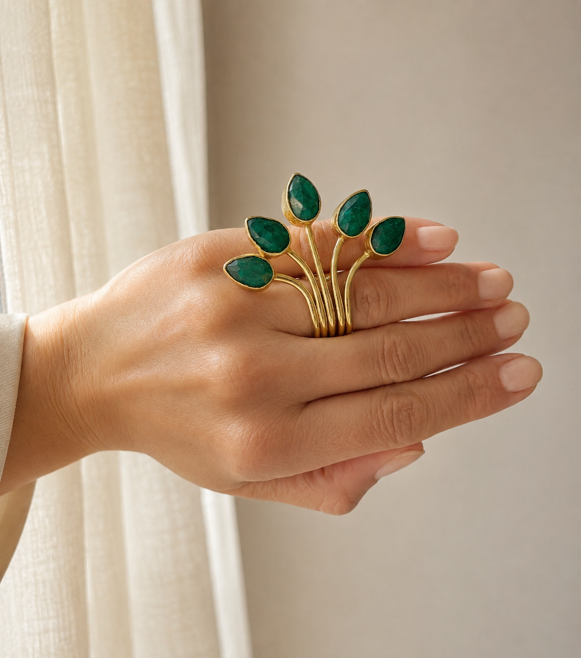 A woman’s hand wearing A gold-plated fan multi-stone emerald ring with five leaf-like branches, shown against a neutral background.